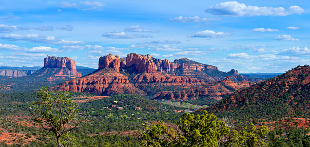 Panoramic view to red rocks near Sedona and the Village of Oak Creek, with lush green forests and bushes in front. The rocks are named Cathedral Rock and Castle Rock.