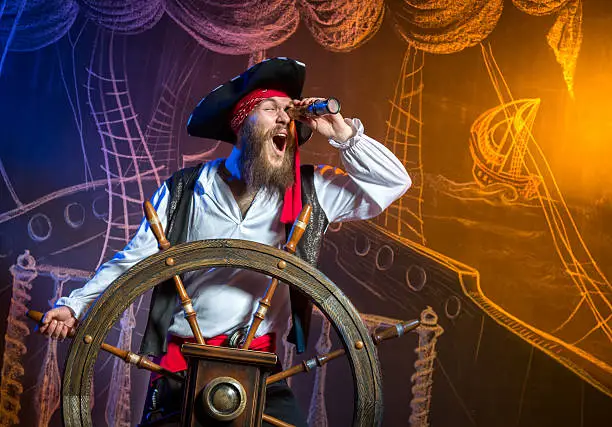 Amusing filibuster standing at the helm of a pirate ship and looking through a spyglass. Chalk drawing of a pirate ship floating in the sea on the background