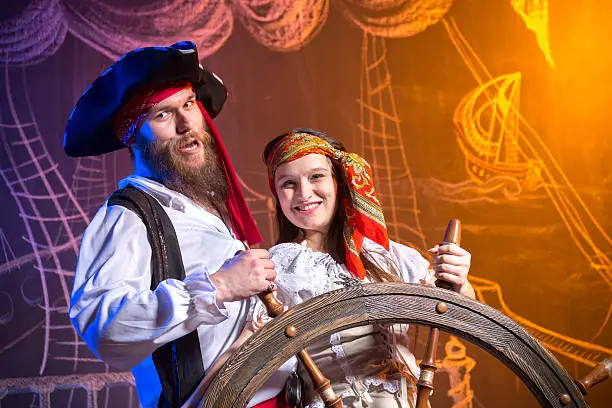 Cheerful couple of pirates at the steering helm of sea ship. Chalk drawing of a pirate ship floating in the sea in the background