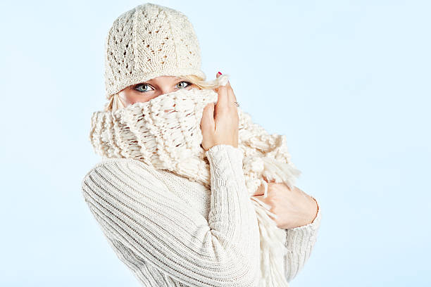 Photo of a young woman in soft sweater, knit cap and scarf, posing against a bright blue background; peeking from behind the scarf wrapped around her face.