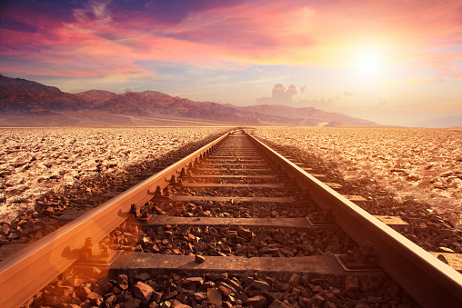 Close-up at ground level of a railroad track running to the horizon in a empty desert at sunset, with sun shining with lens flare. Dramatic sky with cloudscape: red, brown and orange hues. Mountains are far and distant. Arid climate with gravel stones. Straight direction to the way forward. Vaninshing point and personal perspective in the middle of the tracks.
