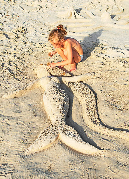 An adorable preschool age girl of Pacific Islander descent stands on the beach, drawing in the sand while spending a leisure day with family outside in Hawaii.