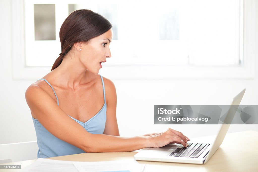 Pretty young woman looking shocked while studying Pretty young woman looking shocked while studying on her laptop in the house desk 2015 Stock Photo