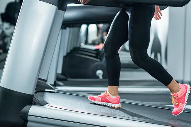 Woman using incline threadmill in modern gym. Incline threadmills are used to simulate uphill walking or running and deliver additional workout benefits to users. Woman is wearing black yoga pants andrunning sports shoes.