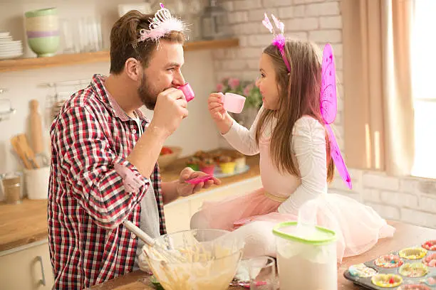 Father and his cute daughter baking and having tea party in kitchen. Sitting and talking, holding toy tea cups. Little girl and father wearing princess costume and tiaras. Table is messy, covered with flour.