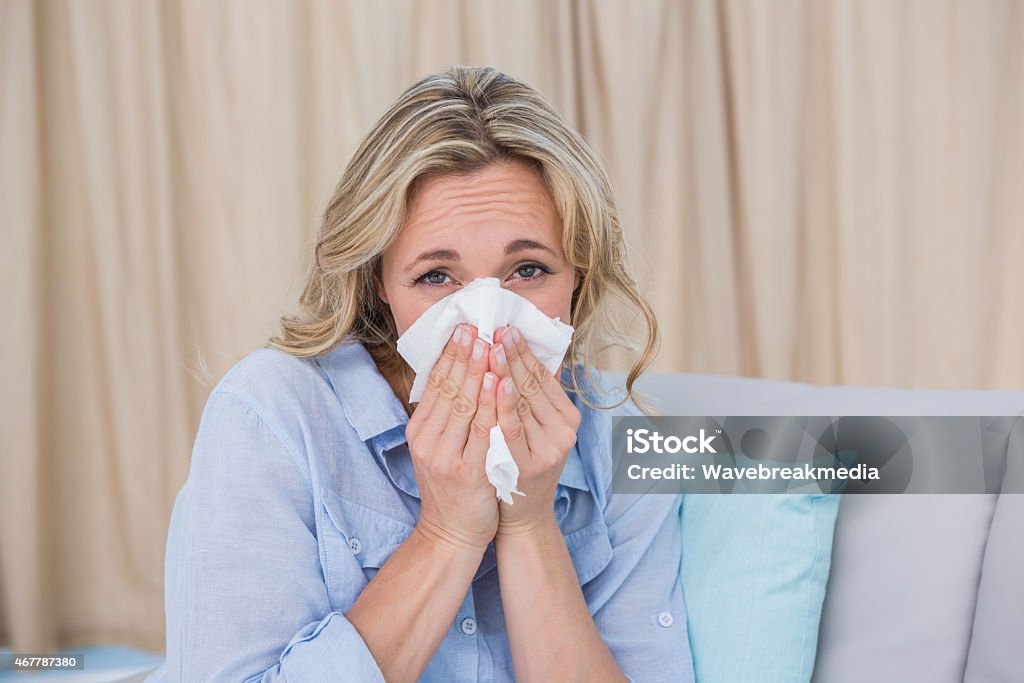 Sick blonde on couch sneezing Sick blonde on couch sneezing on tissue at home in the living room 2015 Stock Photo