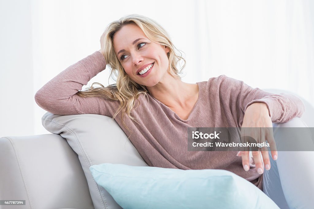 Blonde sitting on couch smiling and thinking Blonde sitting on couch smiling and thinking at home in the living room Mid Adult Women Stock Photo