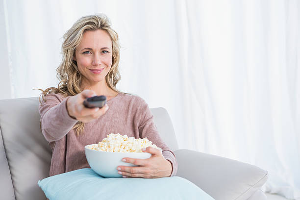 istock Smiling blonde changing tv channel while eating popcorn 467787616