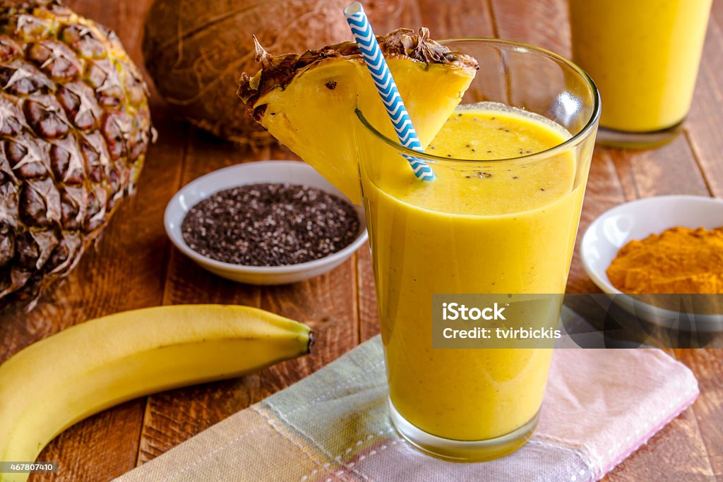 Pineapple, Banana, Coconut, Turmeric and Chia Seed Smoothies Close up fresh blended fruit smoothies made with pineapple, banana, coconut, turmeric and chia seeds surrounded by raw ingredients in drinking glass with pineapple slice garnish and blue striped straw Turmeric Stock Photo