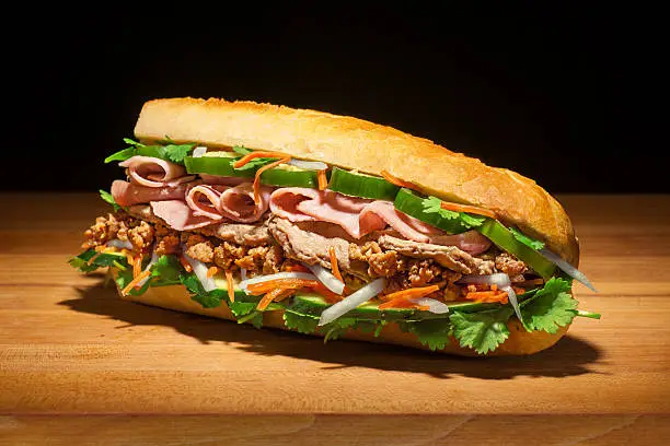 Aspirational style shot of a huge Bánh mì sandwich with cilantro, jalepenos, pickled carrots and daikon, Vietnamese ham, chopped pork and pork steak, and cucumbers, on fresh baguette.