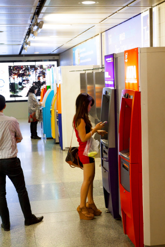 Bangkok, Thailand - February, 3rd 2014: Pretty young Thai woman is standing at ATM in subway and MRT station. She is wearing short red dress, carrying a bag and is using mobile while using ATM. A man is standing at left side. I n background is another woman standing at another ATM. At right are are many ATM different banks.