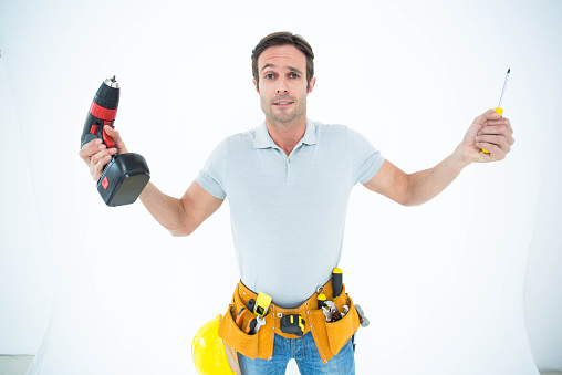 Portrait of confused technician holding screwdriver and drill machine over white background