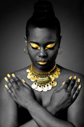 Beautiful African tribal fashion with gold eyeshadow and necklace on dark with arms crossed and looking down.