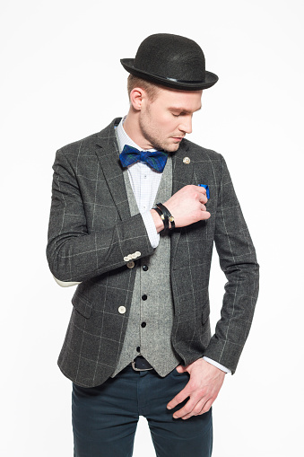 Portrait of elegant blonde young businessman wearing tweed vest, jacket, bow tie and bowler. Standing against white background. Studio shot, one person. 
