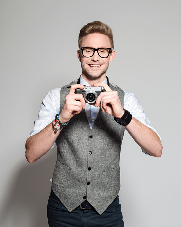 Portrait of happy blonde young photographer wearing tweed vest, bow tie and nerd glasses. Standing with hand in pocket, holding camera and smiling at camera. Studio shot, grey backgound, one person. 