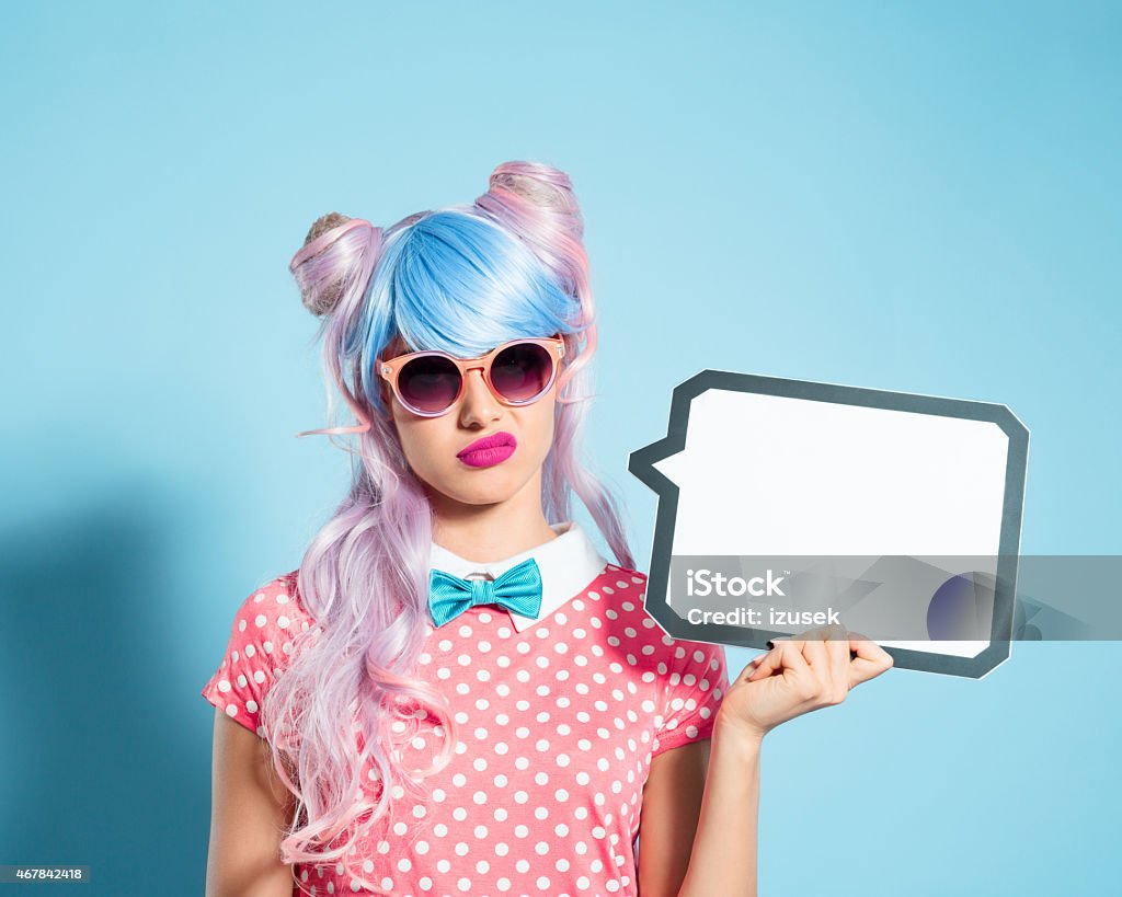 Pink hair manga style girl holding speech bubble Portrait of grimacing manga style blue-pink hair girl wearing sunglasses and pink polka dot dress with collar and bow tie. Standing against blue background, holding a speech bubble in hand. Studio shot, one person. Disgust Stock Photo