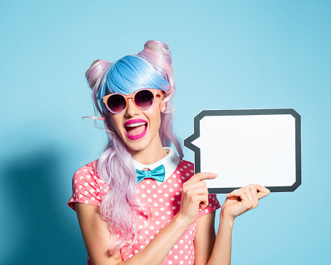 Portrait of excited manga style blue-pink hair girl wearing sunglasses and pink polka dot dress with collar and bow tie. Standing against blue background, holding a speech bubble in hand and laughing at camera. Studio shot, one person.