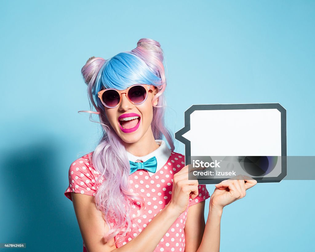 Happy pink hair manga style girl holding speech bubble Portrait of excited manga style blue-pink hair girl wearing sunglasses and pink polka dot dress with collar and bow tie. Standing against blue background, holding a speech bubble in hand and laughing at camera. Studio shot, one person. Advertisement Stock Photo