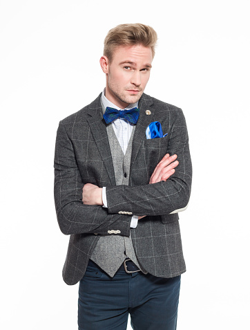 Portrait of elegant blonde young businessman wearing tweed vest, jacket and bow tie. Standing with arms crossed and looking at camera. Studio shot, white backgound, one person. 