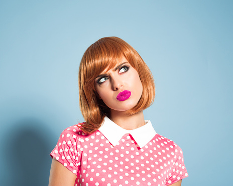 Portrait of thinking red hair young woman wearing polka dot pink dress. Standing against blue backgorund, looking up with doubtful facial expression. Studio shot, one person, headshot.