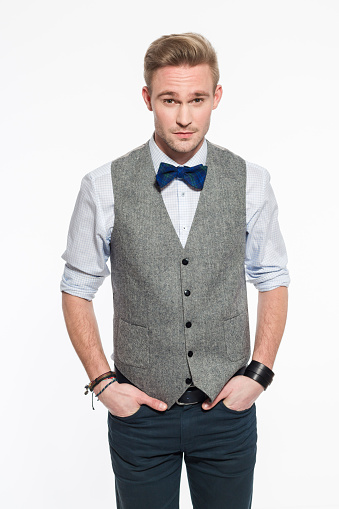 Portrait of confident blonde young businessman wearing tweed vest and bow tie. Standing with hands in pockets and looking at camera. Studio shot, white backgound, one person. 