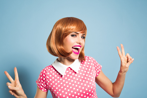 Portrait of happy red hair young woman wearing polka dot pink dress. Standing against blue backgorund, gesturing vicory v sign and laughing at camera. Studio shot, one person, headshot.