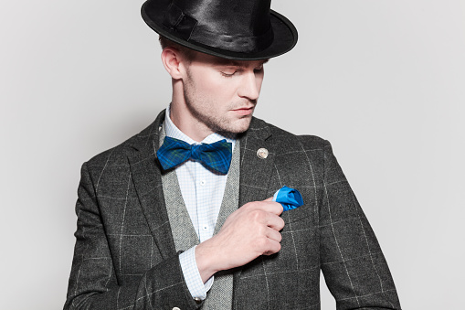 Portrait of elegant blonde young businessman wearing tweed jacket, vest, bow tie and bowler. Standing against grey background. Studio shot, one person. 