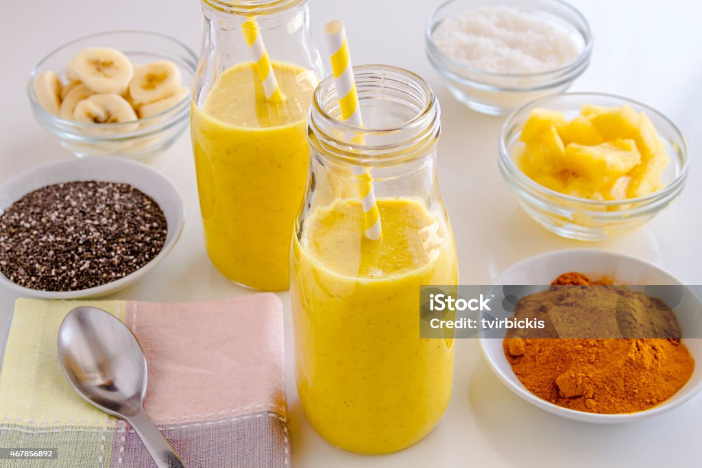 Pineapple, Banana, Coconut, Turmeric and Chia Seed Smoothies Fresh blended fruit smoothies made with pineapple, banana, coconut, turmeric and chia seeds surrounded by raw ingredients yellow straws Pineapple Stock Photo