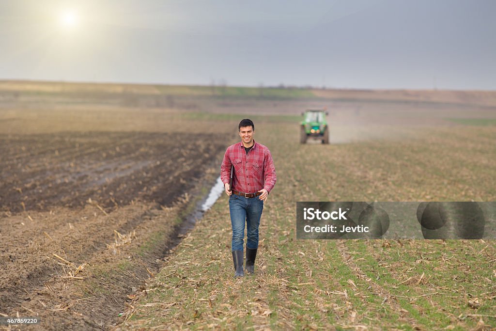 Farmer with laptop and tractor Young landowner with laptop supervising work on farmland, tractor in background Farmer Stock Photo