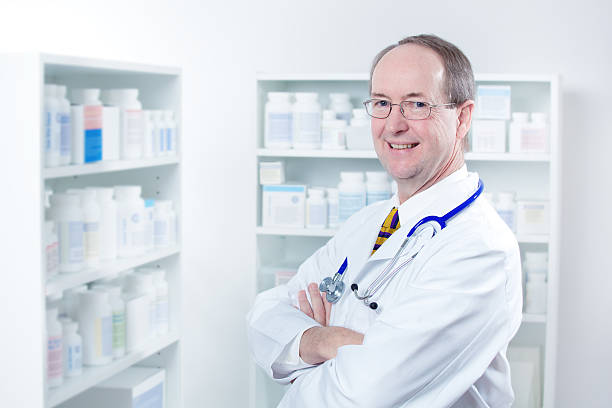 Caucasian Man Pharmacist in Retail Pharmacy Drug Store Horizontal view of a male doctor or medical healthcare professional staff, dressed in a white lab coat, shirt and tie. He stands smiling with his arms folded and a stethoscope around his neck, next to shelves storing pharmaceutical drugs. (No specific information on the bottles is in focus or readable.) suboxone doctor stock pictures, royalty-free photos & images