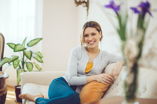 Portrait of a young latin woman who is sitting on a white sofa on sunny day. She is smiling confidently at camera. She is wearing blue jeans, yellow blouse and gray sweater.