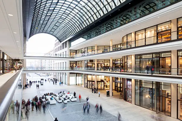 Modern shopping mall with outdoor seating and three floors of stores. There are motioned blurred people sitting, shopping and walking around. The architecture is modern and elegant.