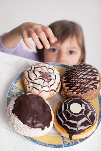 Little girl trying to take one cake from the table. On the table is a plate with four donuts. Little boy is waiting that girl give him one donuts.