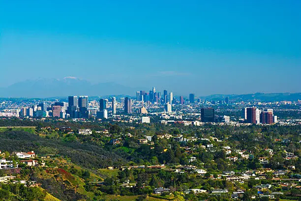 Los Angeles city overview with houses on the foothills of Pacific Palisades and Brentwood in the foreground, West Los Angeles / Westside skylines of Century City, Westwood, and Brentwood midway, and Downtown Los Angeles and Mount Baldy and the San Gabriel Mountains in the background.