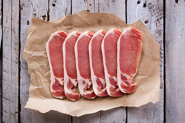 Bacon on Wood On overhead image of a group of bacon slices, on top of baking paper, on rustic worn wood, shot using natural daylight. uncooked bacon stock pictures, royalty-free photos & images