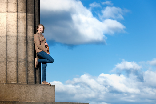 Scottish young woman enjoying sun at the national monument of Scotland in Edinburgh. Woman dressed in a traditional tweed jacket. Leaning against a classical column, sky and cloud in the background