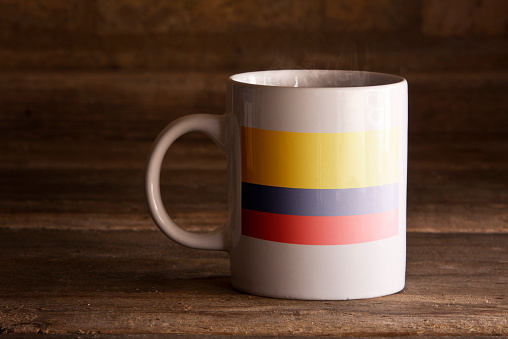 A macro shot of a white ceramic mug with a stenciled flag on the front, shot using natural daylight, on a wooden surface.
