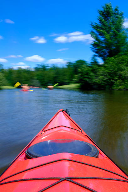 The bow of a red kayak is in sharp focus.  While the environment iti s traveling in is blurred through motion with a long exposure technique.  There are two out of focus kayaks leading the boat from which the image was taken.  Shot with a Fuji X100S and polarizing filter on Michigan's famous AuSable River.