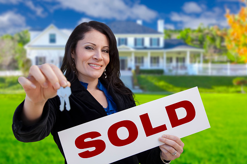 Hispanic Woman Holding Sold Sign and Keys In Front of Beautiful House.
