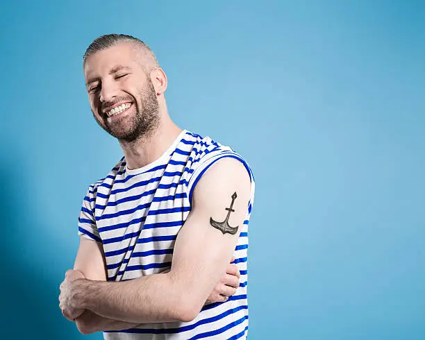 Portrait of happy bearded sailor man with anchor tatoo on shoulder wearing white and blue striped clothing. Standing with arms crossed against blue background and laughing with eyes closed. Studio shot, one person.