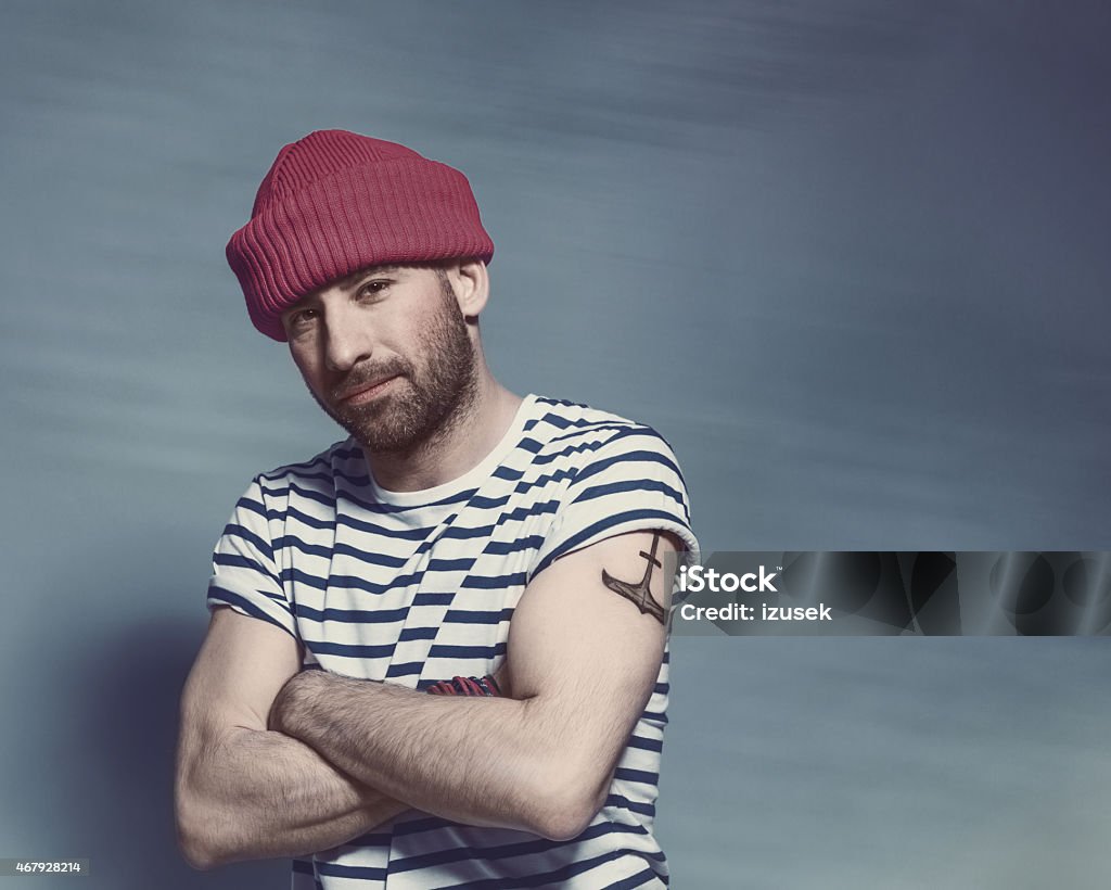 Bearded sailor man wearing striped t-shirt and wool cap Portrait of confident bearded sailor man with anchor tatoo on shoulder wearing white and blue striped clothing and red wool cap. Standing with arms crossed against blue background, looking at camera. Studio shot, one person. Colored Background Stock Photo