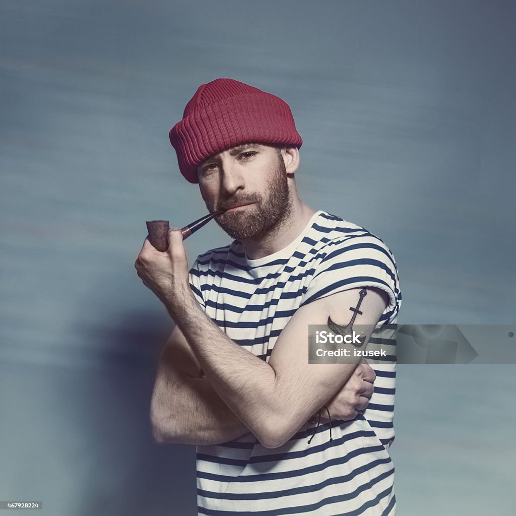Bearded sailor man smorking pipe Portrait of confident bearded sailor man with anchor tatoo on shoulder wearing white and blue striped clothing and red wool cap. Standing against blue background, looking at camera and smoking pipe. Studio shot, one person. Sailor Stock Photo