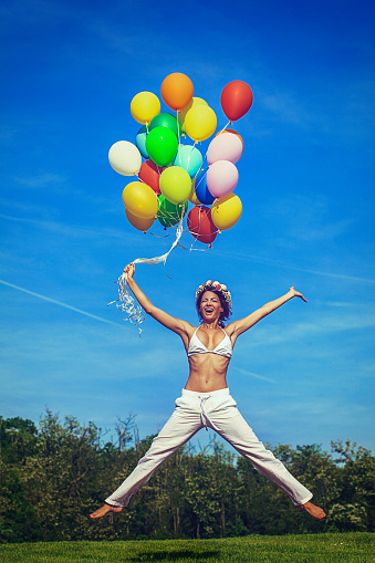 A happy young woman holding colorful balloons, jumping and flying in the air across the green meadow. The woman's mouth, arms and legs are wide spread. She wears white bikini/top tank, yoga pants and a flower wreath in the hair. The blue sky with wispy clouds is in the background; Developed from RAW; Retouched with special care and attention; Small amount of grain added for best final impression; Ready made for print and web use.
