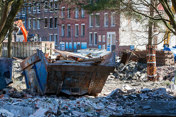 Demolition of residential district Crooswijk in Rotterdam, the Netherlands. Thereâs a lot of rubble in and around a builders skip. The trees are protected by wood against heavy machinery. It looks a bit like a warzone.
