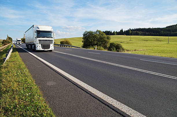 Asphalt road in a rural landscape. The arriving two white trucks on the road. Meadow and forest in the background.