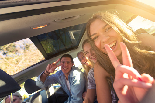 Group of young people in a car. They are off on a road trip and are all happy and smiling and the closest to the camera is making a peace sign. The group are young and could be teenagers or college students. He car has a sunroof and there is some copy space on the left hand side.