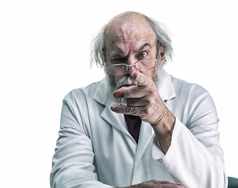 An angry, tangled hair research scientist is pointing his index finger at the viewer. With a stern expression on his face, and one eyebrow raised, he stares over his eyeglasses directly at the camera. He is a balding senior adult man with a gray beard and mustache. He is wearing a white lab coat over his casual polo shirt. Canon 5D Mark III.