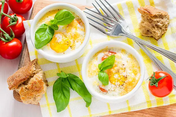 Egg cocotte with cherry tomatoes in white ramekins, bread on plaid napkin, white wood background, top view
