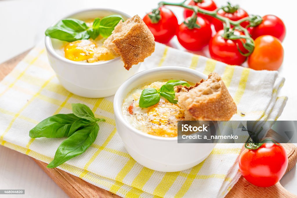 Egg cocotte with cherry tomatoes in white ramekins, bread Egg cocotte with cherry tomatoes in white ramekins, bread on plaid napkin, white wood background Animal Egg Stock Photo