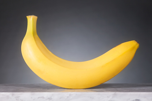 A Banana is laying in front of a gray background on a gray marble plate. Graduated light on the background